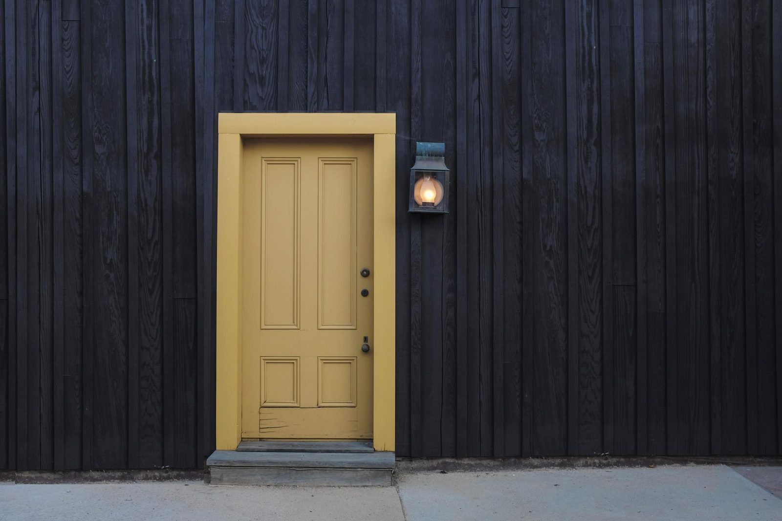 A yellow front door with a lit porch lantern on a dark timber-clad house